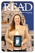 Representative Kristin Bahner is pictured in front of a backdrop of the Minnesota State Capitol while holding the electronic book Out of Africa by Isak Dinesen.  The word READ is spelled out in all capital letters at the top of the image.